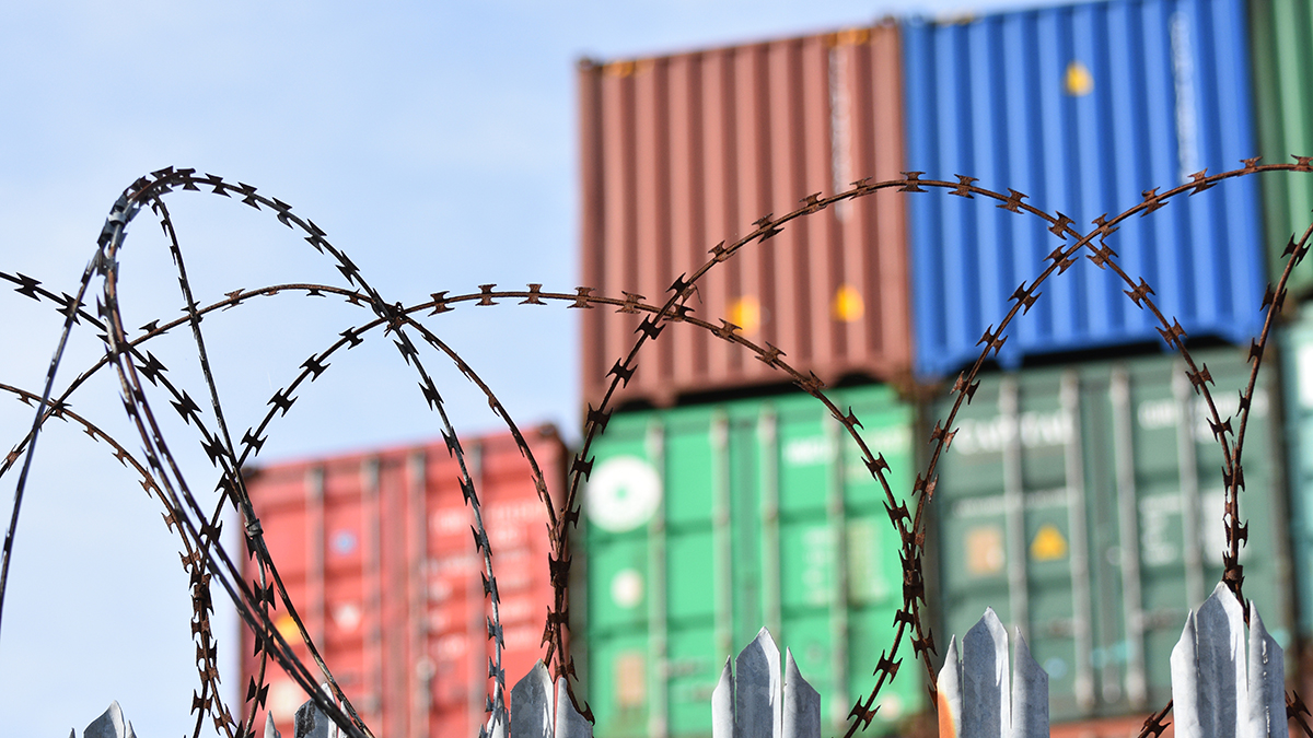 Barbed razor wire in deliberate focus in front of out of focus shipping containers. The image is a metaphor for trade sanctions, embargo, tariffs, hard borders, trade barriers.